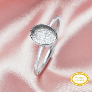 May include: A silver ring with an oval setting. The ring is made of solid gold, as indicated by the text "14K/18K Solid Gold" in a yellow oval. The ring's band is thin and smooth, and the setting has a textured surface.