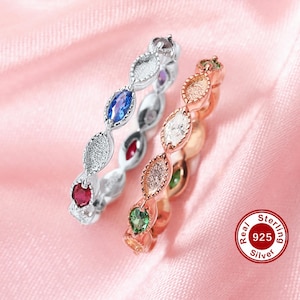 May include: Two rings with colorful gemstones. One ring is silver with red, blue, and purple stones. The other ring is rose gold with green, white, and clear stones. The rings are on a pink satin background.