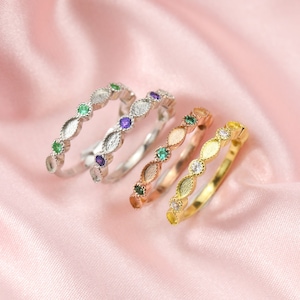 May include: Four dainty rings with oval-shaped settings, each ring features a different colored gemstone. The rings are arranged in a row on a pink satin background. The rings are made of silver, rose gold, and gold.