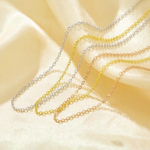 May include: Three delicate chain necklaces in silver, gold, and rose gold. The chains are layered on top of each other and are displayed on a cream-colored fabric background.