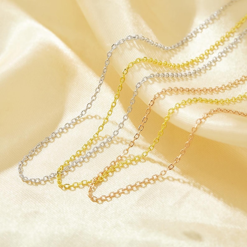 May include: Three delicate chain necklaces in silver, gold, and rose gold. The chains are layered on top of each other and are displayed on a cream-colored fabric background.