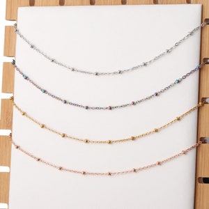 May include: Five different chain necklaces displayed on a wooden background. The chains are silver, rainbow, gold, rose gold, and silver. Each chain has small round beads.