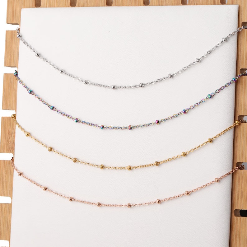 May include: Five different chain necklaces displayed on a wooden background. The chains are silver, rainbow, gold, rose gold, and silver. Each chain has small round beads.