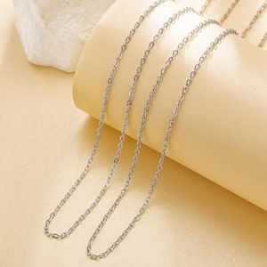 May include: Several silver-colored chain necklaces with a delicate, linked design. The chains are displayed on a light beige surface, creating a simple and elegant presentation. The necklaces appear to be made of metal.