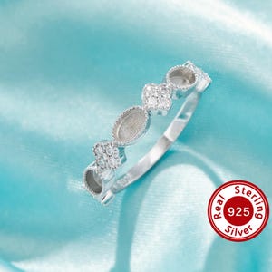 May include: A silver ring with an alternating pattern of round and oval settings, interspersed with small, sparkling stones. The ring is made of sterling silver, as indicated by the red seal with the text "Real Sterling 925 Silver". The ring is displayed on a light blue satin surface.