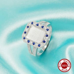 May include: A silver ring with a square-shaped setting surrounded by blue gemstones. The ring has a plain band and is designed to hold a personalized inscription.
