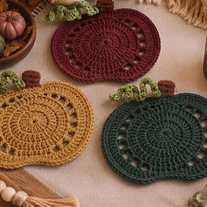 May include: Three round, flat, crocheted pumpkin-shaped coasters in deep burgundy, mustard yellow, and dark forest green. Each coaster features a central circular pattern, an open-work border, a small brown stem, and a curly green vine detail.