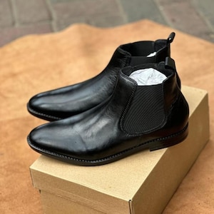 Può includere: Un paio di stivaletti Chelsea in pelle nera con finitura liscia, punte arrotondate e pannelli laterali elastici neri. Gli stivaletti hanno un tacco basso e una linguetta sul retro della caviglia, appoggiati su una scatola di cartone marrone.