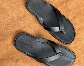 May include: A pair of black leather flip-flops featuring a thong-style strap and a stitched border along the edge of the footbed. The heel area of each sandal is embossed with the text FUTURE FEET PREMIUM FOOT WEAR in a clear, sans-serif font.