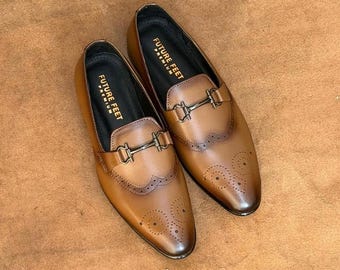 May include: A pair of tan leather loafers featuring a dark metal horsebit detail across the vamp, a wingtip toe cap with decorative brogue perforations, and a black interior lining with the text FUTURE FEET PREMIUM printed in gold lettering.