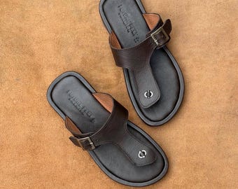 May include: A pair of dark brown leather sandals featuring a T-strap design with an adjustable metal buckle on the side. The footbed is embossed with the text FUTURE FEET and a small metal circular accent is visible on the toe post area.