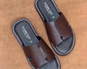 May include: A pair of dark brown slides featuring a textured crocodile-embossed pattern on the wide strap. The footbed is dark brown with white stitching along the edge and the text FUTURE FEET PREMIUM 42 printed in white on the heel area.