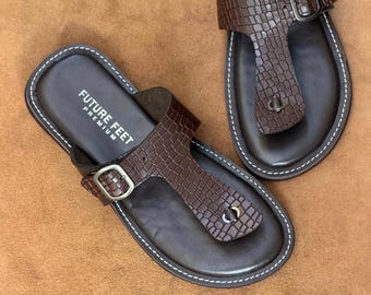 May include: A pair of dark brown thong sandals featuring a crocodile-embossed leather strap with a silver-toned metal buckle. The footbed is smooth dark brown leather with white contrast stitching along the edge and the text FUTURE FEET PREMIUM.
