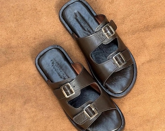 May include: A pair of dark brown leather sandals featuring two wide straps across the top, each secured with a silver-toned metal buckle. The footbed is smooth and dark brown, with a matching dark brown sole and a rounded toe design.