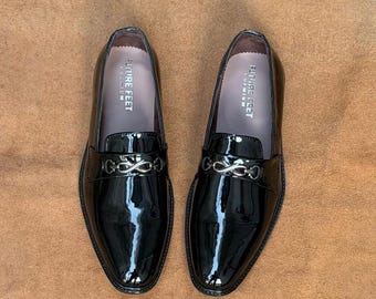 May include: A pair of black patent leather loafers featuring a shiny finish and a silver-toned metal bit detail across the vamp. The interior of each shoe is lined in a light gray material with the text FUTURE FEET PREMIUM printed in white.