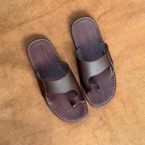 May include: A pair of dark brown leather toe-loop sandals featuring a wide strap across the midfoot and a loop for the big toe. The footbed is stamped with the text FUTURE FEET PREMIUM FOOTWEAR and the edges are finished with white stitching.