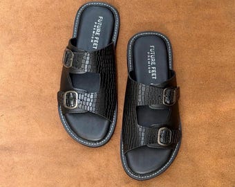 May include: A pair of black sandals featuring a crocodile-embossed texture on the two wide straps. Each strap is secured with a dark metal buckle. The footbed is black with white stitching along the edge and the text FUTURE FEET PREMIUM printed.