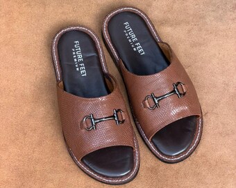 May include: A pair of brown open-toe slides featuring a textured snakeskin-like pattern on the upper strap, a dark brown footbed with the text FUTURE FEET PREMIUM, and a metallic horsebit detail. The edges are finished with white contrast stitching.