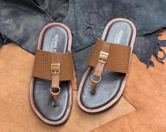 May include: A pair of brown thong sandals featuring a textured crocodile-embossed strap with a gold-tone metal buckle and a central vertical strap. The footbed is dark brown with the text FUTURE PREMIUM printed in white on the heel area.