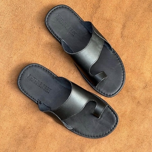 May include: A pair of black leather sandals featuring a toe loop design and a wide strap across the top. The footbed is embossed with the text FUTURE FEET PREMIUM FOOTWEAR. The sandals have visible stitching along the edges of the sole and strap.