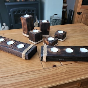 May include: A selection of dark brown wooden candle holders, some rectangular with rope detailing, others square or cylindrical. Each holds a tea light candle. The holders are arranged on a wooden table.