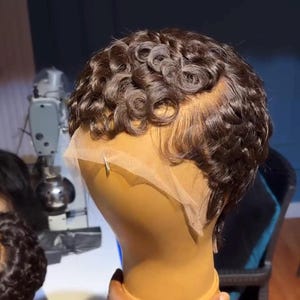 May include: A brown, short-hair wig with tight curls, styled on a mannequin head. The wig features a lace front for a natural hairline. The mannequin is held by a person's hand.