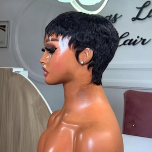 Op de afbeelding: Een mannequinhoofd met een korte, zwarte, gelaagde pruik. De pruik heeft een zijscheiding en is licht golvend. De mannequin heeft volledige make-up, inclusief donkere oogschaduw en dikke wenkbrauwen. Een kleine, ronde, zilveren oorbel is zichtbaar.