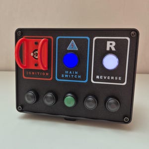 Op de afbeelding: Een zwart bedieningspaneel met verlichte knoppen in rood, blauw en wit. Het paneel heeft een rode contactsleutel, een blauwe hoofdschakelaar en een witte achteruitschakelaar. Extra zwarte en groene knoppen bevinden zich eronder.
