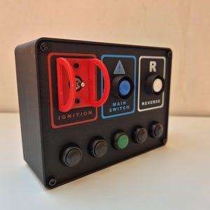 Op de afbeelding: Zwart bedieningspaneel met rode "IGNITION" schakelaar, blauwe "MAIN SWITCH" knop en witte "REVERSE" knop. Inclusief diverse zwarte en groene knoppen. Het paneel is met schroeven bevestigd.