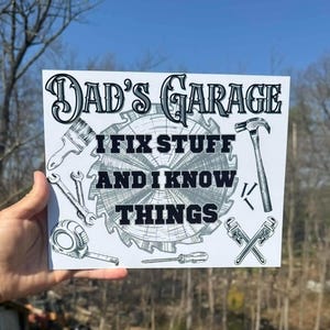 May include: A white sign with the text "Dad's Garage" and the phrase "I fix stuff and I know things." The sign features illustrations of tools like a hammer, wrench, and saw blade. The sign is held up against a background of trees and a blue sky.