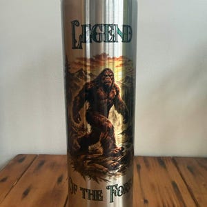 May include: A stainless steel tumbler with a clear lid. The tumbler features a Bigfoot illustration in a forest setting, with the words "LEGEND OF THE FOREST" in a stylized font. The design is printed on the metallic surface.