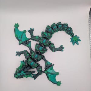 May include: An articulated dragon figurine featuring a dark charcoal gray body with vibrant green accents. The surface is textured with a repeating leaf pattern, and the creature has two wings, a long segmented tail, and a detailed head.