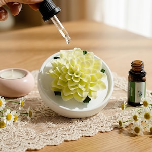 May include: A white ceramic essential oil diffuser with a yellow flower design. A dropper dispenses oil onto the flower. A small brown bottle of jasmine oil and a pink candle holder are also present.