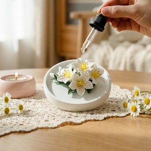 Puede incluir: Un difusor de aromaterapia de cerámica blanca con detalles de flores blancas y amarillas. Una mano usa un cuentagotas para agregar líquido al difusor. Una vela rosa encendida y margaritas también están sobre la mesa.