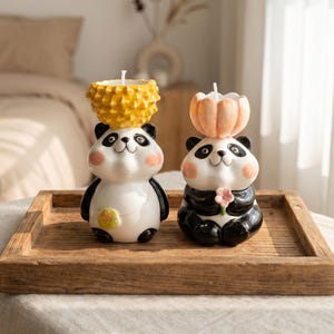 May include: Two panda-shaped candles with black and white bodies, pink cheeks, and smiling faces. One panda has a yellow pineapple-shaped candle on its head, the other has a peach-colored pumpkin candle. They sit on a wooden tray.