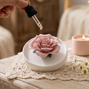 Può includere: Un diffusore di oli essenziali a forma di rosa rosa in una ciotola di ceramica bianca. Un contagocce eroga olio sulla rosa. Sono visibili anche una candela rosa accesa e un centrino all'uncinetto.