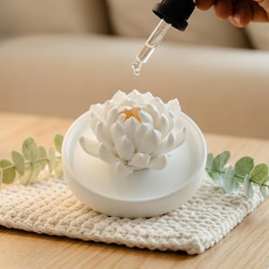 May include: A white ceramic essential oil diffuser shaped like a lotus flower, with a dropper dispensing liquid. The diffuser sits on a cream-colored, crocheted coaster. The flower has a yellow center.