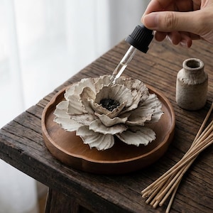 Op de afbeelding: Een bloemvormige diffuser voor etherische olie op een houten dienblad. Een hand gebruikt een druppelaar om olie aan de bloem toe te voegen. Een kleine keramische fles en een bundel riet liggen ook op de houten tafel.