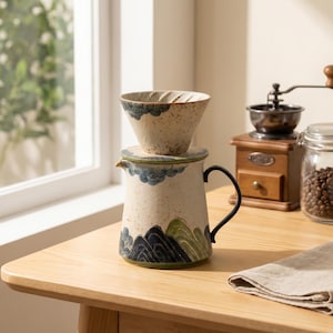 Könnte beinhalten: Ein Keramik-Kaffeefilter-Set mit einer gesprenkelten cremefarbenen Basis und handgemalten blauen und grünen Berg- und Wolkenmotiven. Das Set besteht aus einem konischen Filteraufsatz auf einer passenden Kanne mit schwarzem Metallgriff.