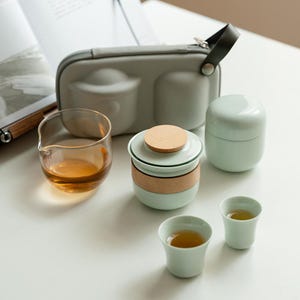 Puede incluir: Un juego de té con una tetera de cerámica verde claro, dos tazas pequeñas y una jarra de vidrio llena de té. También se incluye un estuche de transporte verde claro y un recipiente pequeño. El juego de té está sobre una superficie blanca.