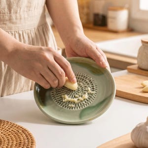 May include: A round, light green ceramic grater plate with a textured, raised centre for grating garlic. The plate features a smooth, glossy finish with darker green marbled accents along the edges, designed for manual food preparation.