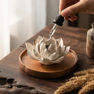 May include: A white lotus flower-shaped essential oil diffuser sits on a wooden tray. A dropper dispenses liquid into the flower's petals. The diffuser is on a dark wooden surface, with a small ceramic bottle in the background.