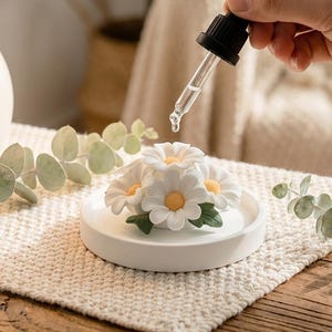 Può includere: Un diffusore di fiori di margherita in ceramica bianca con un contagocce nero che eroga liquido. Il diffusore presenta diversi fiori di margherita bianchi con centri gialli e foglie verdi.