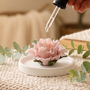 Könnte beinhalten: Ein rosa, blütenförmiger Diffusor für ätherische Öle auf einem weißen Keramiktablett. Eine Glaspipette gibt Flüssigkeit auf die Blüte ab. Grüne Blätter umgeben den Diffusor, vor einem neutralen Hintergrund.