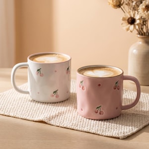Puede incluir: Dos tazas de cerámica, una blanca y otra rosa claro, con un patrón repetitivo de pequeñas cerezas rojas y puntos. Ambas tazas tienen una superficie redondeada y ligeramente texturizada, y contienen café con un diseño de espuma en forma de corazón.