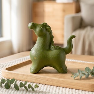 Może przedstawiać: Zielona ceramiczna figurka konia o teksturowanej powierzchni. Koń ma zakrzywiony ogon i szczegółową grzywę. Figurka stoi na drewnianej tacy, element dekoracyjny.
