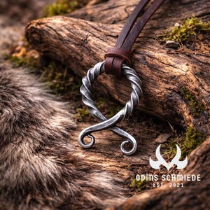 Könnte beinhalten: Ein silberfarbener Anhänger mit gedrehtem Design und braunem Lederband. Der Anhänger hat eine kreisförmige Oberseite und eine stilisierte, geschwungene Unterseite. Der Hintergrund zeigt einen Holzstamm und Fell.