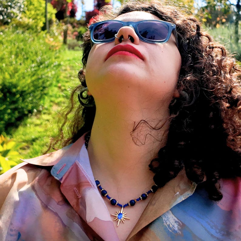 Worn outdoors in sunlight, the blue strand sits at the base of the neck and the gold starburst pendant rests just below the collarbone against an open pastel collar.