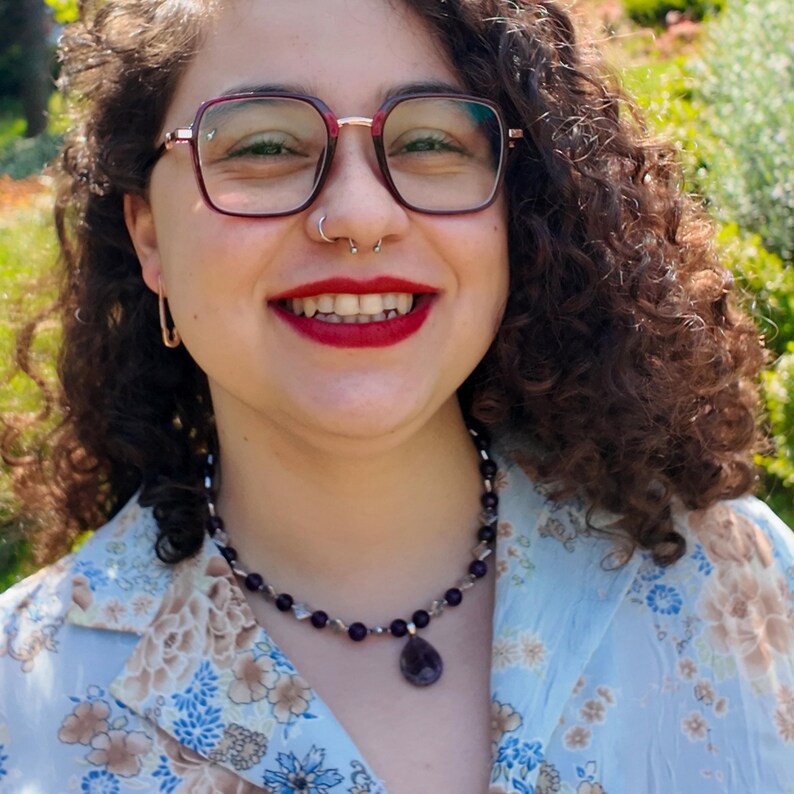 Front view in sunlight: the strand of round purple beads circles the base of the neck over an open floral shirt collar, the teardrop pendant hanging centred a short drop below the beads.