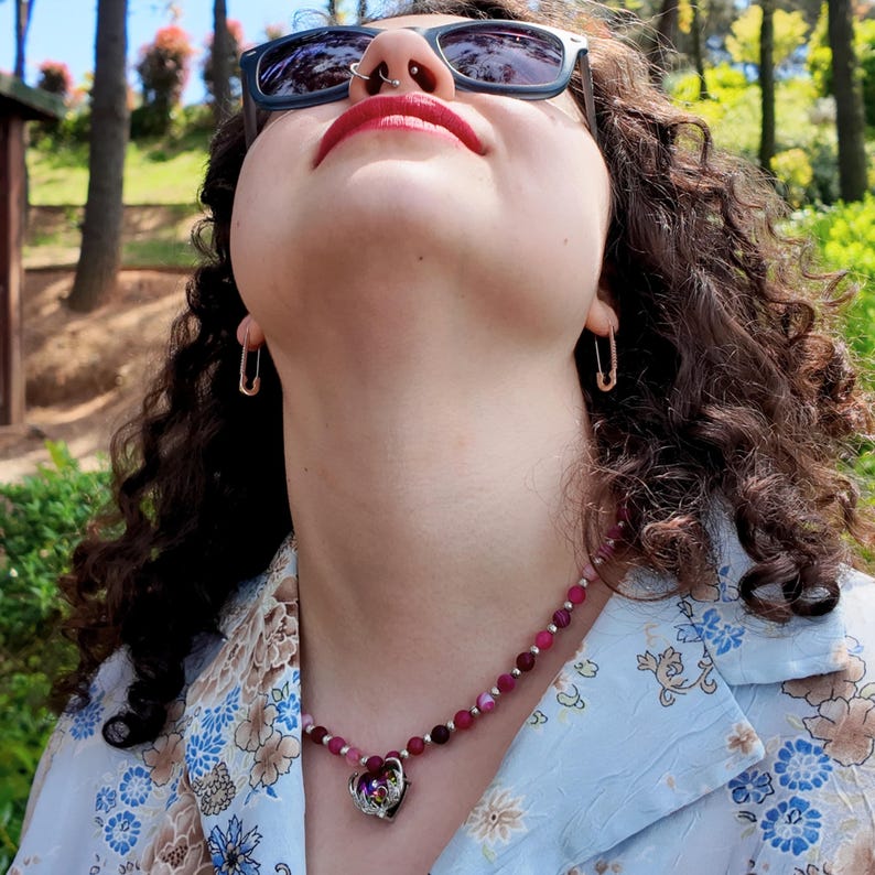 Close daylight view against skin: fuchsia, wine and pale banded beads alternating with tiny bright metal rounds, finishing in a heart pendant whose stone throws violet and green light.